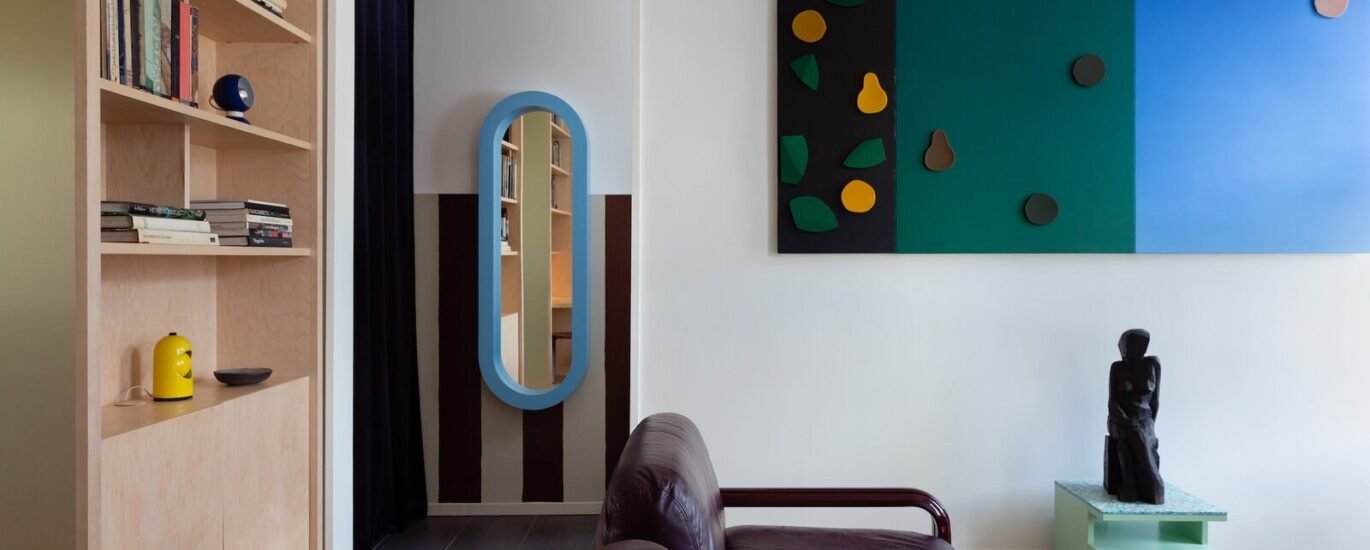 A stylish living room interior featuring a brown leather sofa with wooden arms, a light wood built-in bookshelf filled with books and decorative objects, a blue oval-framed full-length mirror in a doorway with dark curtains, a colourful geometric wall art piece in teal, black, and blue with fruit-sh