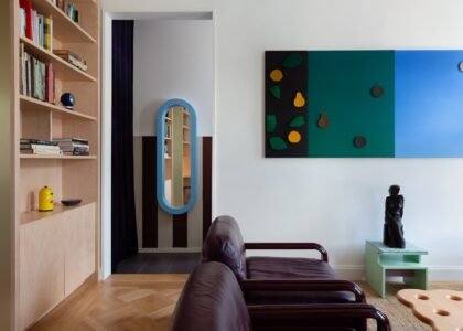 A stylish living room interior featuring a brown leather sofa with wooden arms, a light wood built-in bookshelf filled with books and decorative objects, a blue oval-framed full-length mirror in a doorway with dark curtains, a colourful geometric wall art piece in teal, black, and blue with fruit-sh