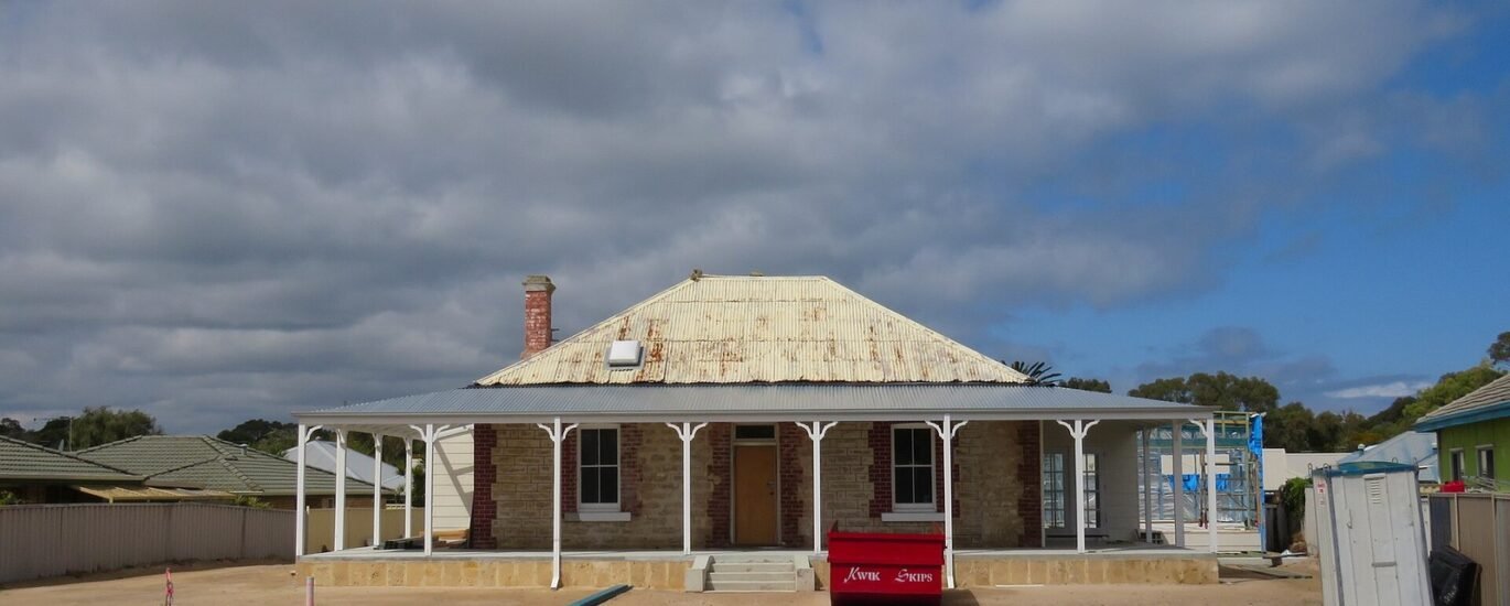 A heritage-listed single-storey stone cottage with a corrugated iron hip roof, brick chimney and white verandah posts, surrounded by a bare dirt yard with a red skip bin and construction materials in the foreground, under a cloudy sky in Rockingham, Western Australia, during renovation works.