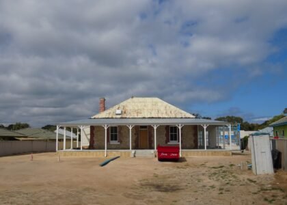 A heritage-listed single-storey stone cottage with a corrugated iron hip roof, brick chimney and white verandah posts, surrounded by a bare dirt yard with a red skip bin and construction materials in the foreground, under a cloudy sky in Rockingham, Western Australia, during renovation works.